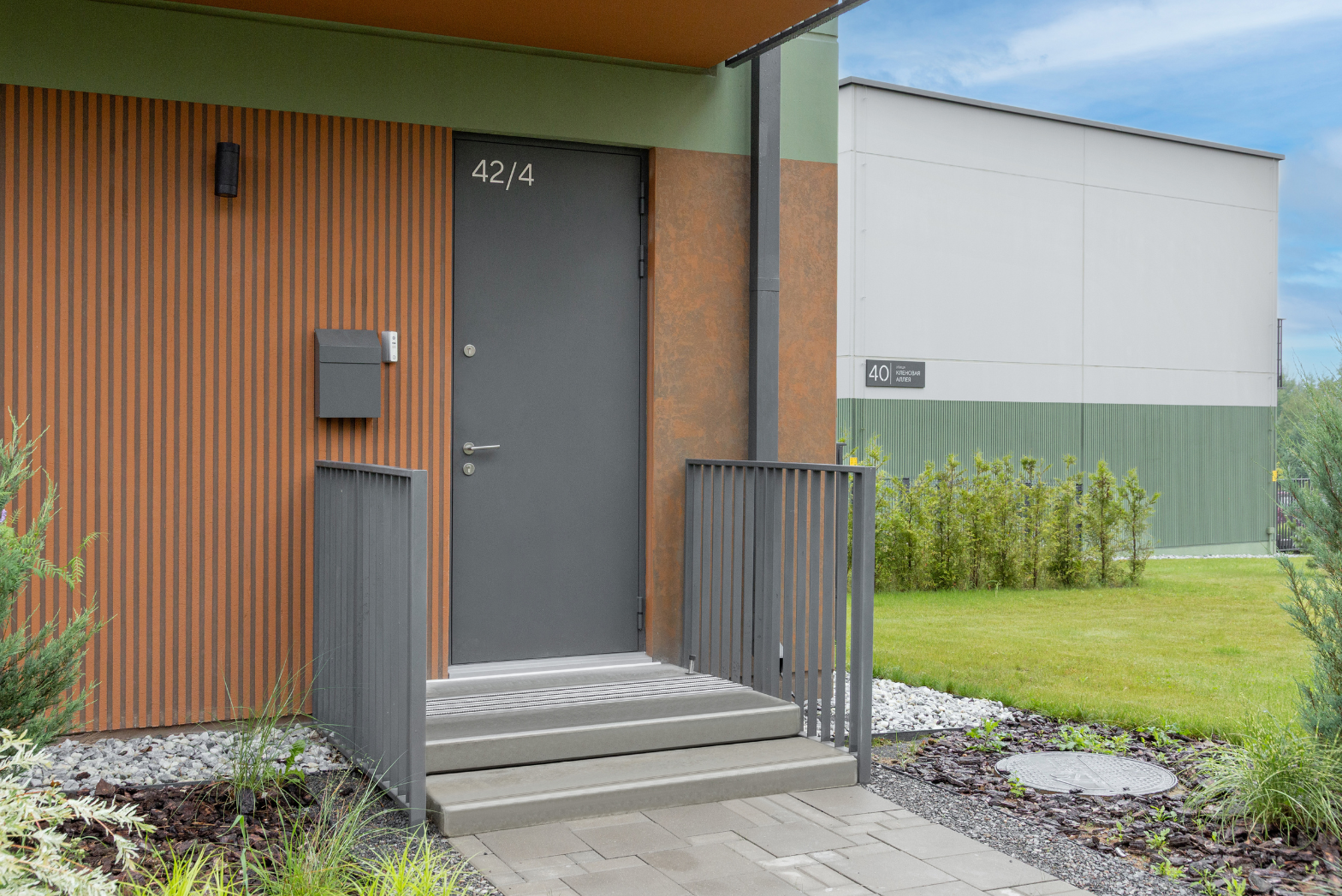 Входные двери для дома тёмно-серого цвета, на фоне деревянной облицовки, со ступеньками и перилами.